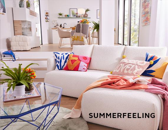 Ein gemütliches Wohnzimmer mit einem weißen Sofa, das mit bunten Kissen dekoriert ist. Ein moderner Couchtisch und Pflanzen runden die sommerliche Atmosphäre ab. Der Text "SUMMERFEELING" ist auf dem Bild zu sehen.