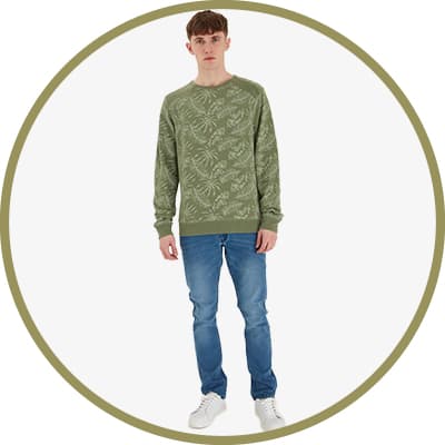 Mann in hellgrünem Pullover und Jeans steht lächelnd vor einfarbigem Hintergrund