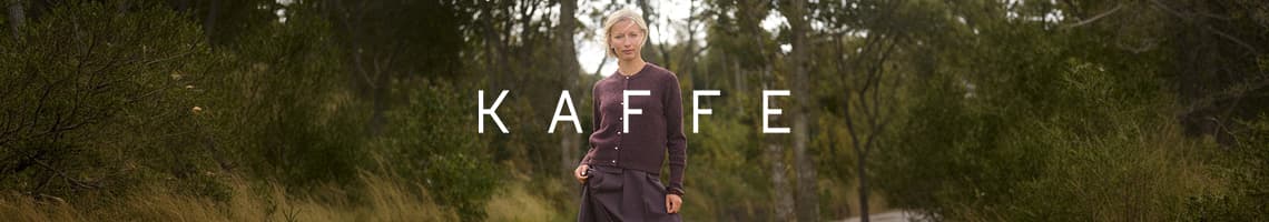 KAFFE – Person im dunklen Cardigan und Rock steht auf Waldweg zwischen hohen Büschen
