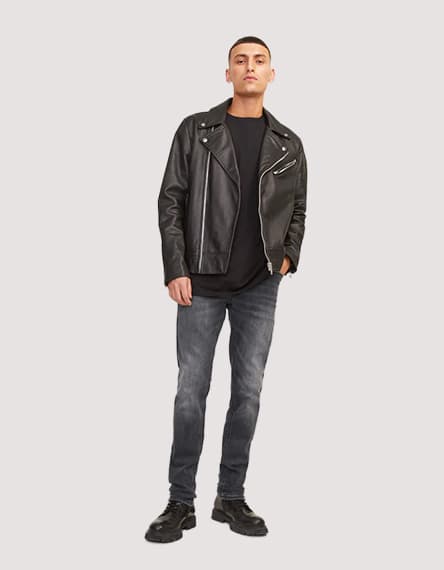Mann steht frontal zur Kamera in schwarzer Jacke, dunklem Shirt, grauer Jeans und schwarzen Schnürschuhen