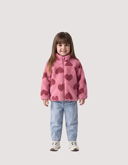 Kind steht frontal in rosa Jacke mit Herzmuster, hellen Jeans und Turnschuhen vor hellem Hintergrund