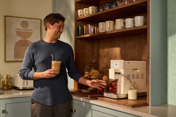 Mann in Küche bereitet mit Kaffeevollautomat ein Getränk zu und hält ein Glas Eiskaffee in der Hand