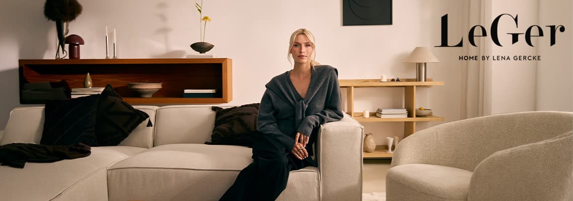 Frau in dunkler Strickjacke sitzt entspannt auf hellem Sofa in modernem Wohnzimmer mit Regalen und Deko im Hintergrund