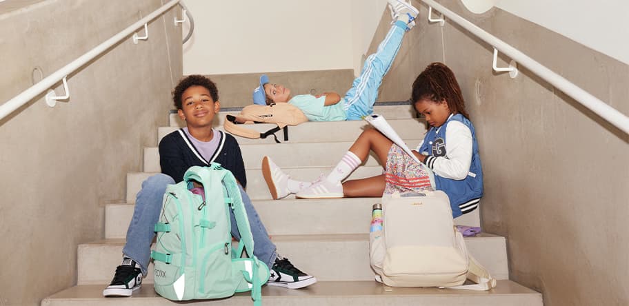 Drei Kinder sitzen und liegen auf Treppe, tragen bunte Freizeitkleidung und haben Rucksäcke. Gesichter sind verschwommen.