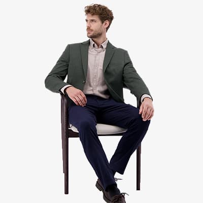 Mann in grünem Sakko und beiger Hemd sitzt auf einem Stuhl, trägt dunkelblaue Hose und schwarze Schuhe.
