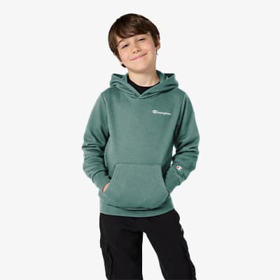 Junge in grünem Kapuzenpullover mit weißen Logos auf der Brust und dem Ärmel, Hände in der Kängurutasche, trägt schwarze Hose