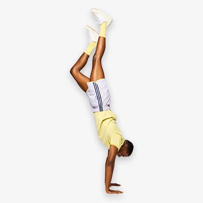 Mann in gelbem T-Shirt und weißen Shorts macht einen Handstand, beide Beine in gelben Socken ausgestreckt nach oben.