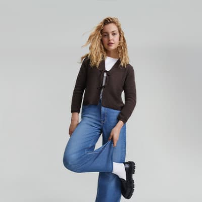 Frau mit langen Haaren trägt braunen Cardigan, weiße Socken und blaue Jeans. Sie posiert mit angewinkeltem Bein.