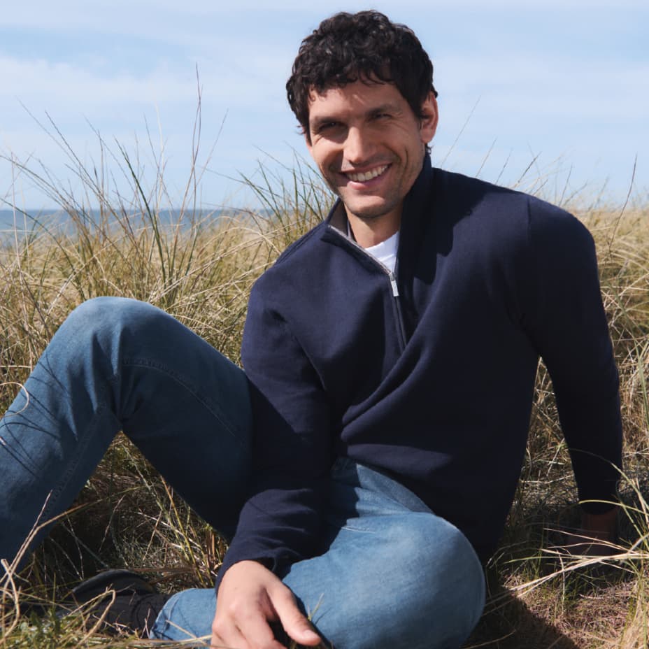 Mann in dunkelblauem Pullover und Jeans sitzt im Gras, Gesicht unscharf, entspannte Körperhaltung, blauer Himmel im Hintergrund