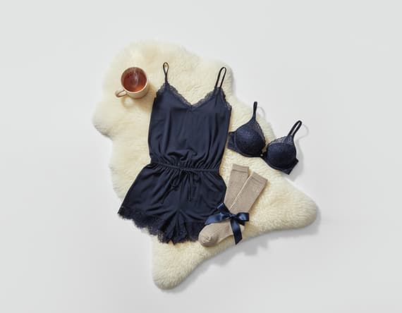 Dunkelblauer Spitzenpyjama, passender BH, beige Socken mit Schleife und Tasse Tee auf weißem Fell.