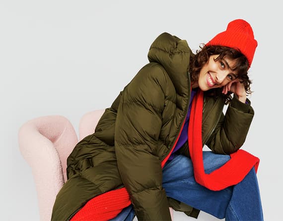 Frau sitzt in rosa Sessel, trägt grünen langen Daunenmantel, rote Mütze und Schal, darunter blaue Jeans und lila Pullover.