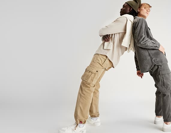 Zwei Personen lehnen sich Rücken an Rücken, tragen lässige Kleidung in Beige und Grau, stehen vor neutralem Hintergrund.