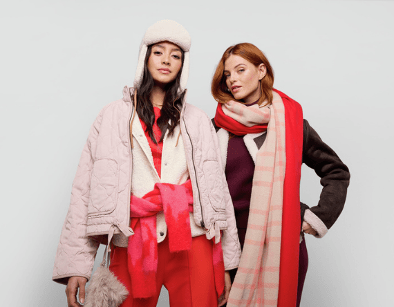 Zwei Frauen in Winterkleidung mit Schals und Jacken posieren nebeneinander vor weißem Hintergrund.