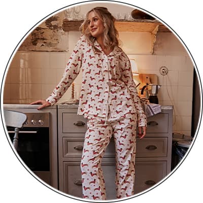 Frau in weißem Pyjama mit roten Hundemotiven lehnt an Küchenarbeitsplatte, Ofen und Kaffeemaschine im Hintergrund