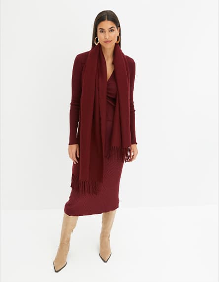 Frau in weinrotem Kleid und passendem Schal, trägt beige Stiefel und goldene Ohrringe, steht vor weißem Hintergrund