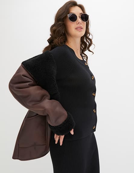 Frau mit welligem Haar trägt schwarzen Strickpullover und braune Jacke über Schulter. Gesicht ist verschwommen.