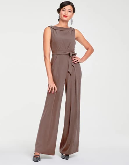 Frau in ärmellosem braunen Jumpsuit mit weitem Bein und Taillengürtel. Sie steht mit einer Hand auf der Hüfte.