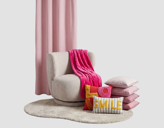 Gemütlicher Sessel mit pinker Decke, gestapelte Kissen und Kissen mit dem Schriftzug "SMILE" vor rosa Vorhang.