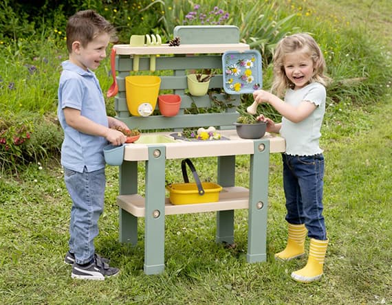 Zwei Kinder spielen im Garten an einem grünen Spieltisch mit Pflanzen und Gartenwerkzeugen.