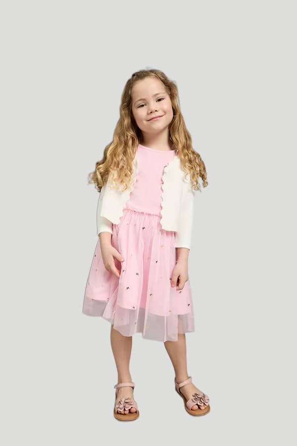 Kind in rosa Kleid und weißer Strickjacke, trägt Sandalen mit Blumenapplikationen.