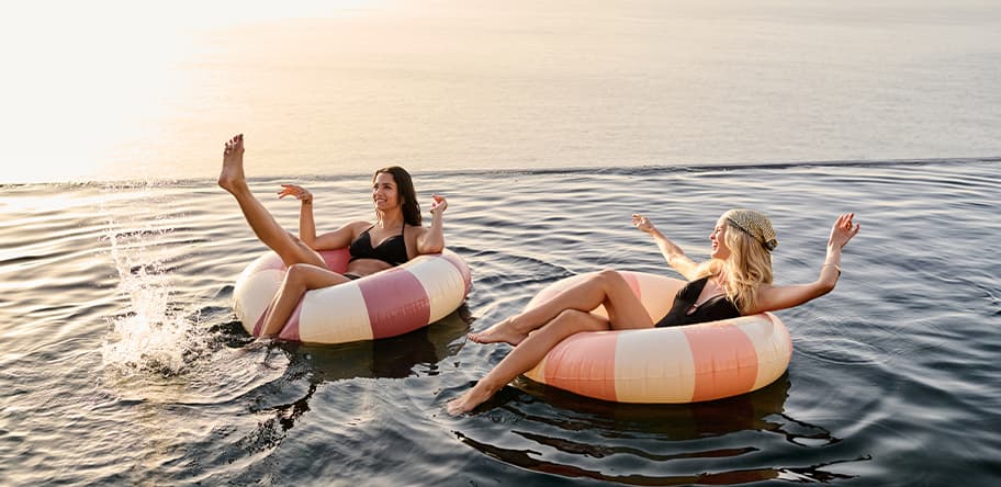 Zwei Frauen in Badeanzügen entspannen auf Schwimmringen im Wasser, eine Frau spritzt mit den Füßen.