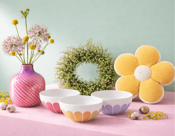 Bunte Schüsseln, Vase mit Blumen, grüner Kranz und gelbes Kissen in Blumenform auf rosa Tisch.