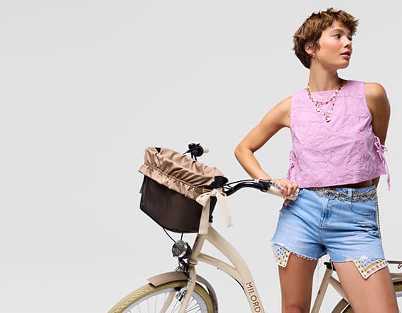 Frau in lila Oberteil und blauen Shorts lehnt lässig an cremefarbenem Fahrrad mit Korb.