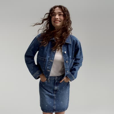 Frau mit welligem Haar trägt Jeansjacke und Jeansrock, beide Hände in den Taschen, vor grauem Hintergrund.