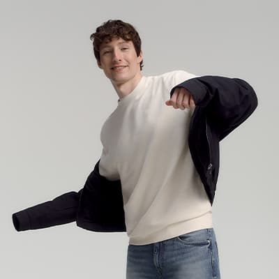 Mann zieht schwarze Jacke über cremefarbenen Pullover an, trägt blaue Jeans.