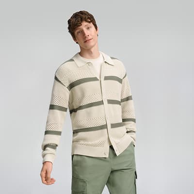 Person in gestreifter Strickjacke und grüner Hose, Hand in der Hosentasche, blickt zur Seite.