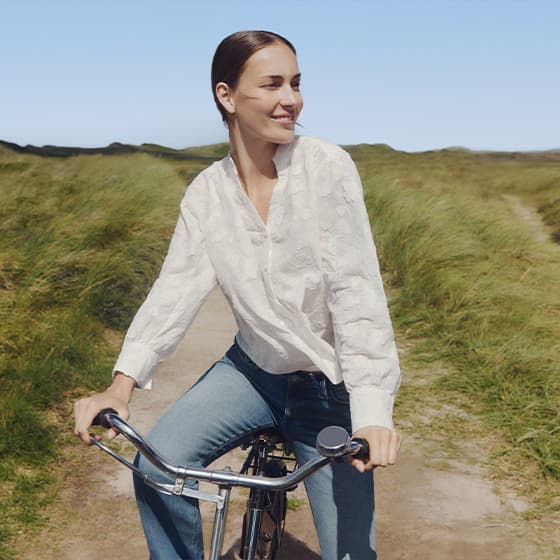 Frau in weißer Bluse und blauer Jeans fährt Fahrrad auf ländlichem Weg.