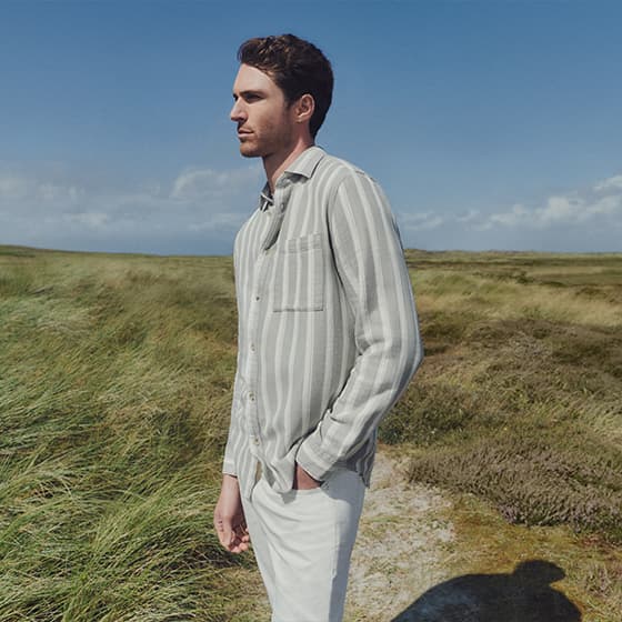 Mann in gestreiftem Hemd und heller Hose steht in einer grünen Landschaft mit blauem Himmel.
