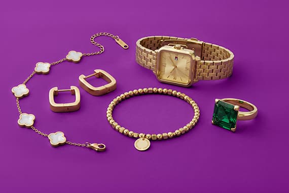 Armbanduhr, Armband, Ring mit grünem Stein, Ohrringe und Blumenarmband liegen auf violettem Hintergrund