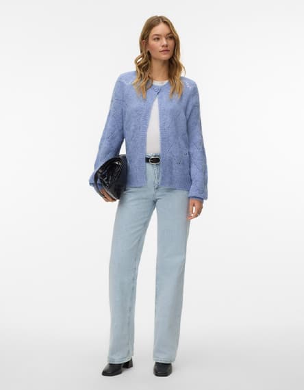 Frau in hellblauer Strickjacke und weißem Top, hellblaue Jeans, schwarze Stiefeletten, hält schwarze Clutch in der Hand