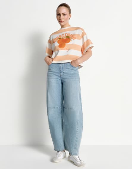 Frau im gestreiften T-Shirt mit Orangenprint und Schriftzug steht in weiter hellblauer Jeans und Sneakers vor weißem Hintergrund