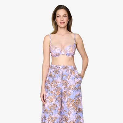 Frau in gemustertem Bikinioberteil und weiter, passender Hose mit Palmblättern steht frontal mit Händen in den Hosentaschen