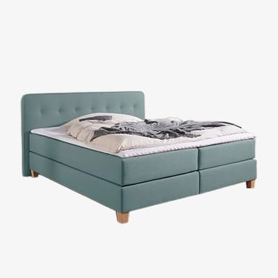 Grünes Boxspringbett mit hohem Kopfteil, weißer Bettdecke und grauer Decke, auf Holzfüßen freistehend
