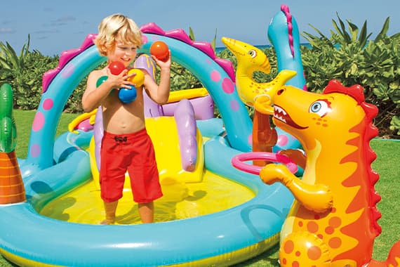 Kind spielt im bunten Dino-Planschbecken mit aufblasbaren Figuren und bunten Bällen im Garten