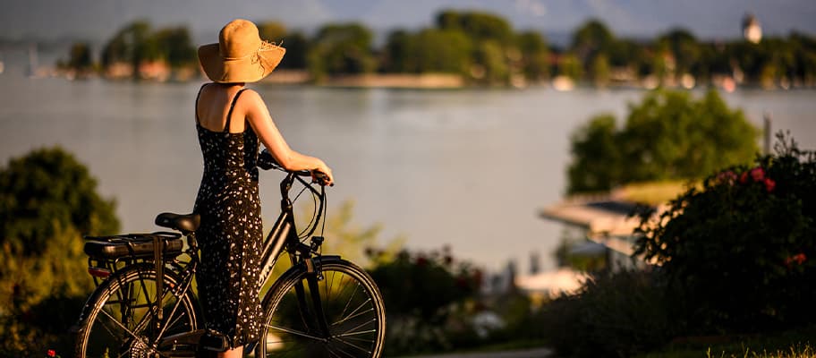 Frau mit Sommerhut steht mit Fahrrad am Seeufer und blickt über das Wasser auf eine grüne Insel