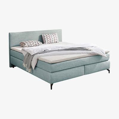 Helles Boxspringbett mit hohem Kopfteil, Decke und zwei gemusterten Kissen, leicht unordentlich aufgelegt