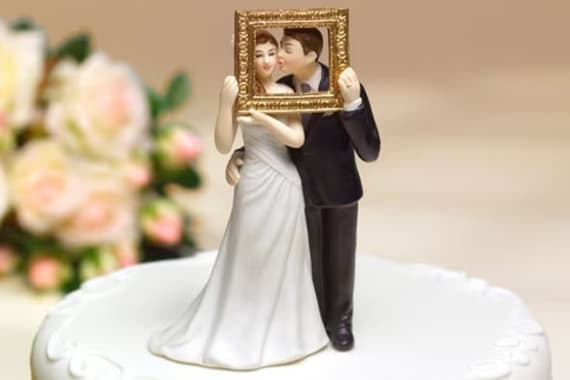 Hochzeitstortenfigur mit Braut und Bräutigam, die gemeinsam einen leeren Bilderrahmen vor ihre Gesichter halten