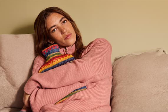 Person auf Sofa umarmt bunte Wärmflasche und trägt weiten rosa Pullover