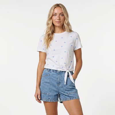 Frau steht frontal und trägt weißes T-Shirt mit Knoten vorn und gemusterter Shorts in Jeansoptik
