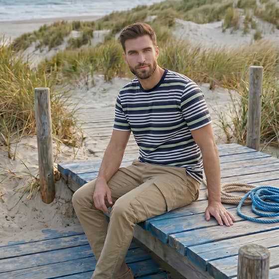 Mann im gestreiften T-Shirt und Chinohose sitzt entspannt auf Holzsteg am Strand mit Dünen im Hintergrund