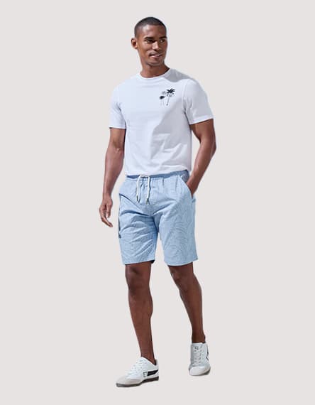 Mann steht in weißem T‑Shirt mit kleinem Palmenmotiv und hellen Shorts mit Kordelzug, eine Hand in der Tasche