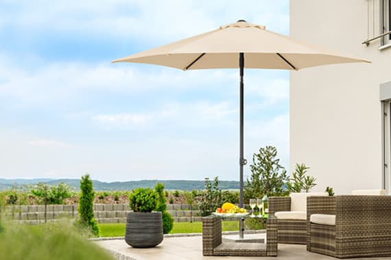 Großer Sonnenschirm auf Terrasse mit Sitzgruppe, Obstschale und Getränken, im Hintergrund Garten und weite Landschaft
