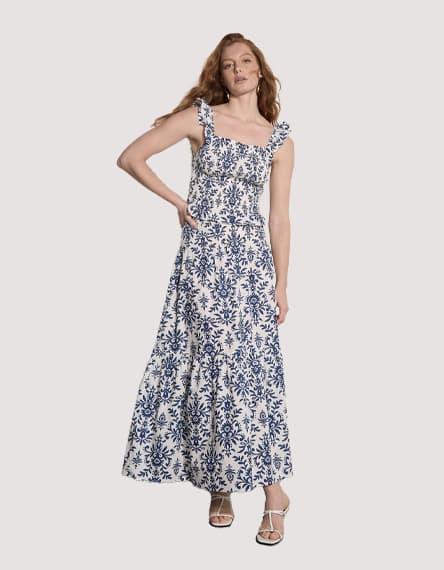 Frau steht in langem weißen Kleid mit blauem Blumenmuster und Trägern, eine Hand in der Hüfte, neutrale Pose