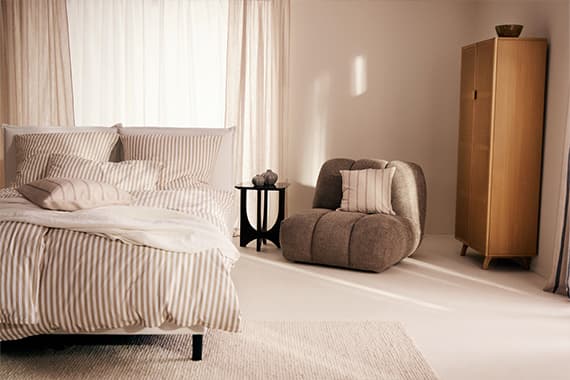 Schlafzimmer mit gestreifter Bettwäsche, weichem Sessel mit Kissen, kleinem Beistelltisch und schmalem Kleiderschrank