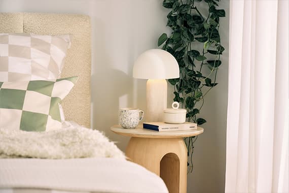 Nachtkästchen mit Lampe, Tasse, Buch und Dose neben Bett mit kariertem Kissen und herabhängender Zimmerpflanze