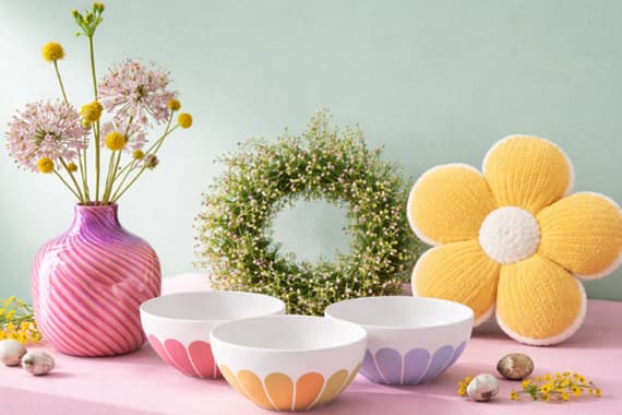 Bunte Schüsseln, Vase mit Blumen, grüner Kranz und gelbes Kissen in Blumenform auf rosa Tisch.